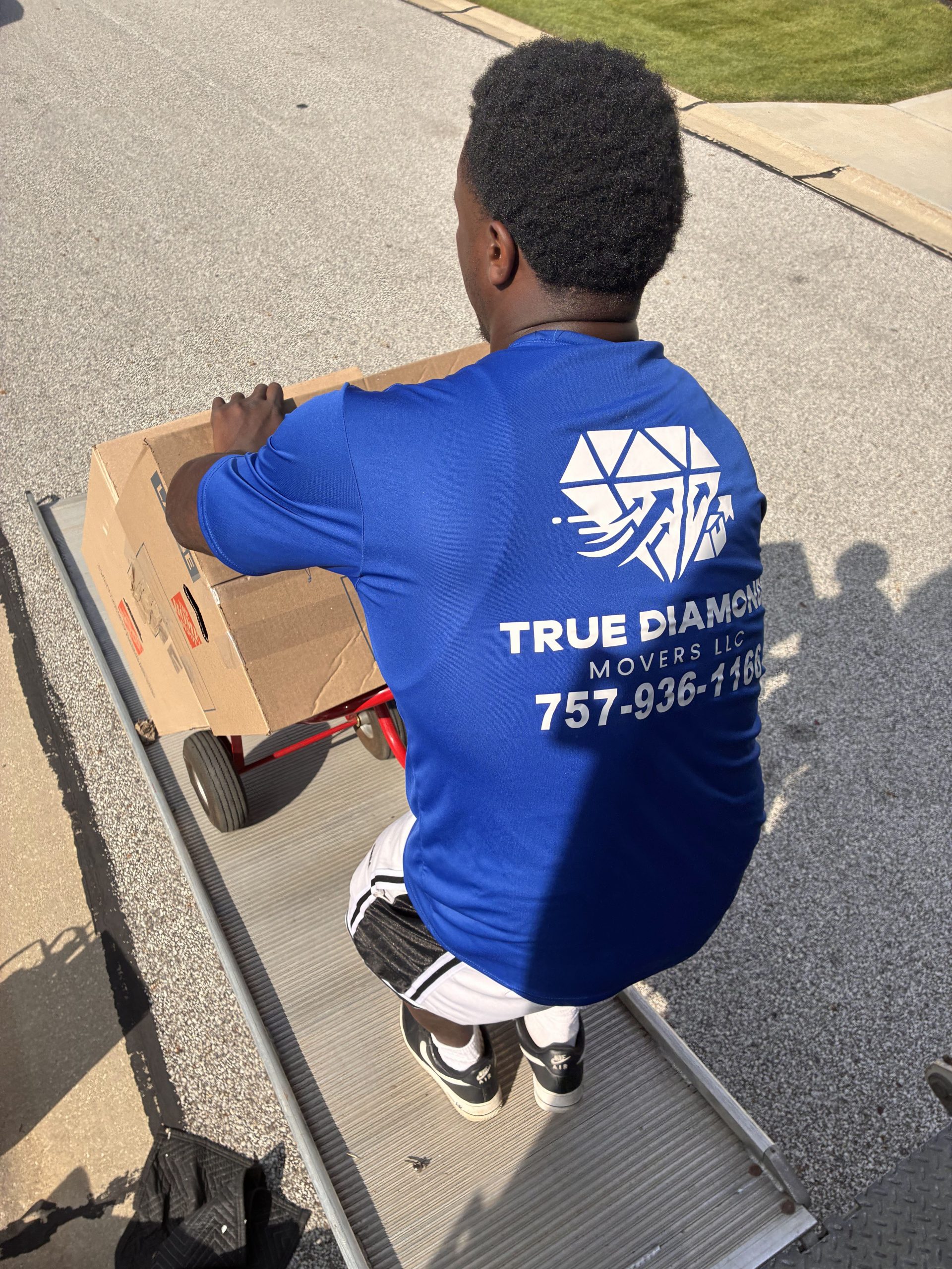 A mover in a blue True Diamond Movers shirt pushes a boxed load on a hand truck along a sunny residential street.