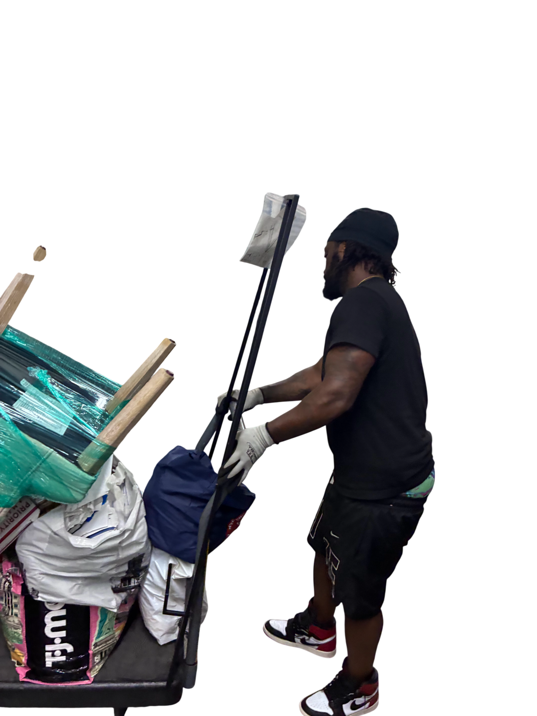 Man wearing gloves and a beanie pushes a cart loaded with bags and wrapped furniture pieces against a black background.