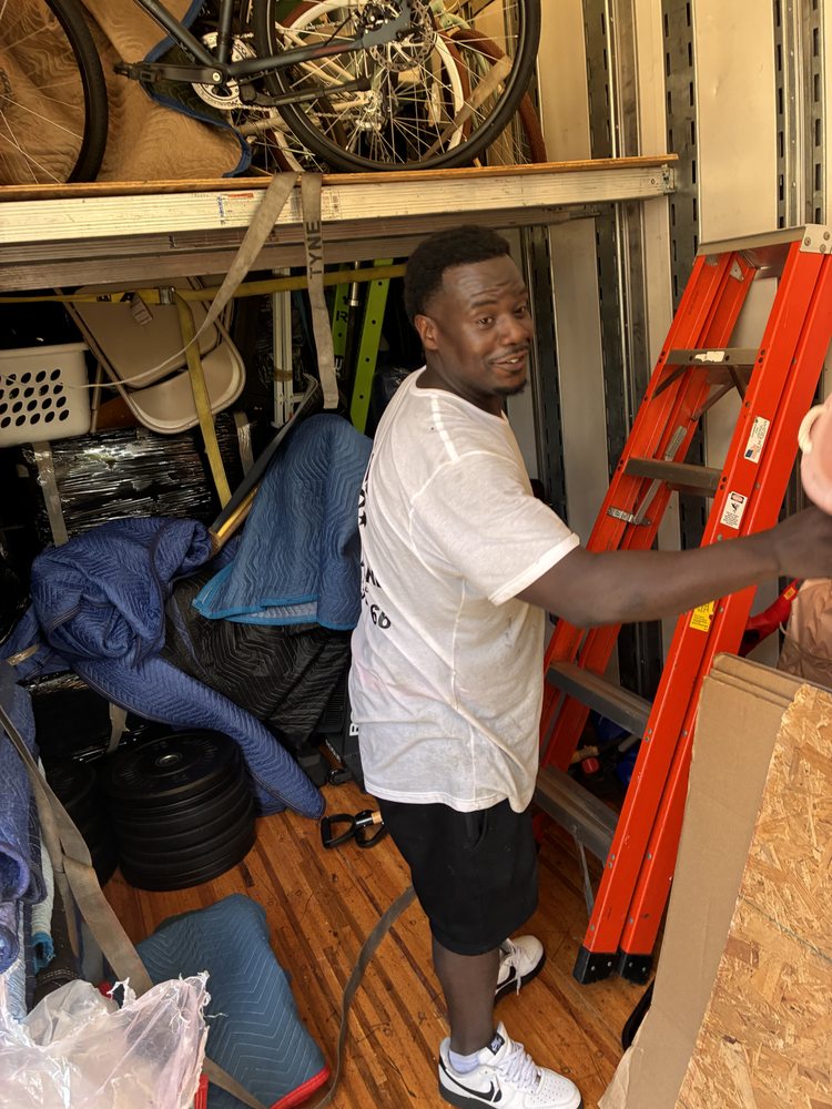 Smiling man in a white shirt stands inside a cluttered storage truck beside an orange ladder, bicycles, furniture, and household items.