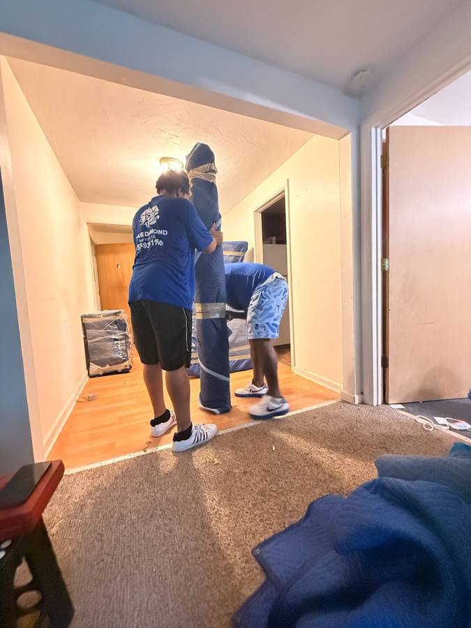Three movers in blue uniforms carry a large couch through a doorway in a warmly lit home, creating a busy, collaborative moving scene.