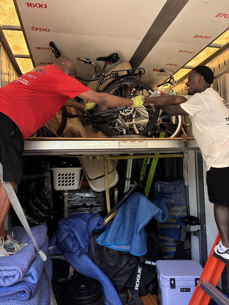 Two men work together to load a bicycle into a moving truck, surrounded by blankets, storage bins, and other bikes; scene conveys teamwork.