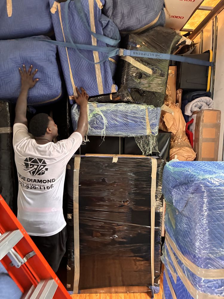 Worker in a moving truck pushes wrapped furniture into a tightly packed load of blue-padded boxes, secured with straps; busy moving scene.